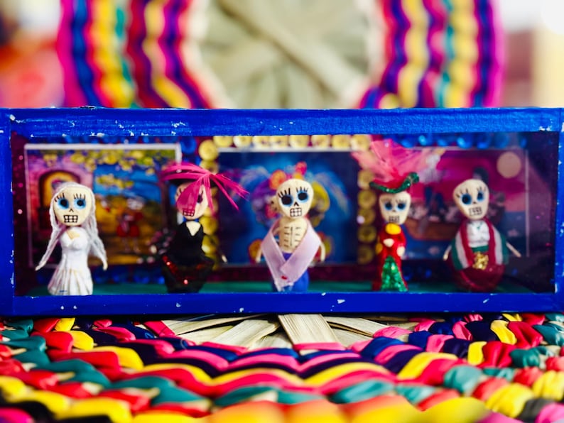 May include: A blue framed display case featuring five Day of the Dead figurines. Each figurine has a skull face and is dressed in colorful attire. The background is a colorful, woven pattern.
