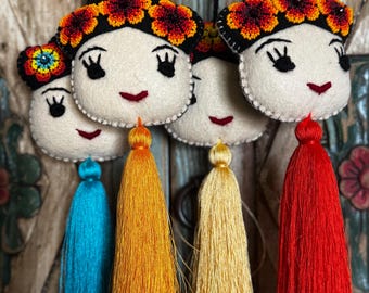 May include: Handmade felt ornaments featuring smiling faces with black hair, floral headbands, and colorful tassels. The tassels are in shades of blue, yellow, and red. Each ornament is suspended by a black string.