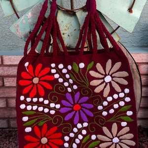 May include: A burgundy tote bag with embroidered floral designs in red, purple, and beige. The bag features braided burgundy handles and white dotted accents. The bag is displayed against a rustic background.