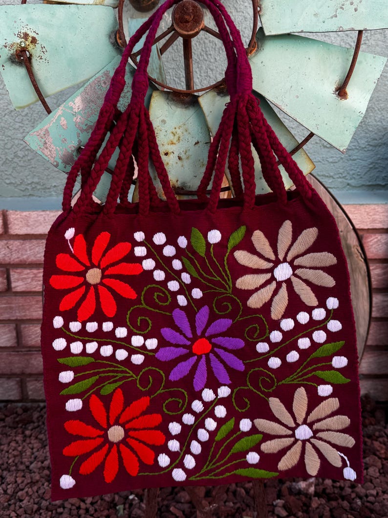 May include: A burgundy tote bag with embroidered floral designs in red, purple, and beige. The bag features braided burgundy handles and white dotted accents. The bag is displayed against a rustic background.