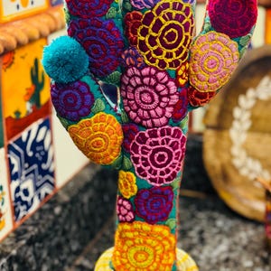 Op de afbeelding: Een kleurrijke geborduurde cactus met pompoenen erop, staand in een geweven pot. De cactus is gemaakt van stof en heeft een verscheidenheid aan felle kleuren, waaronder rood, roze, geel, blauw en groen.