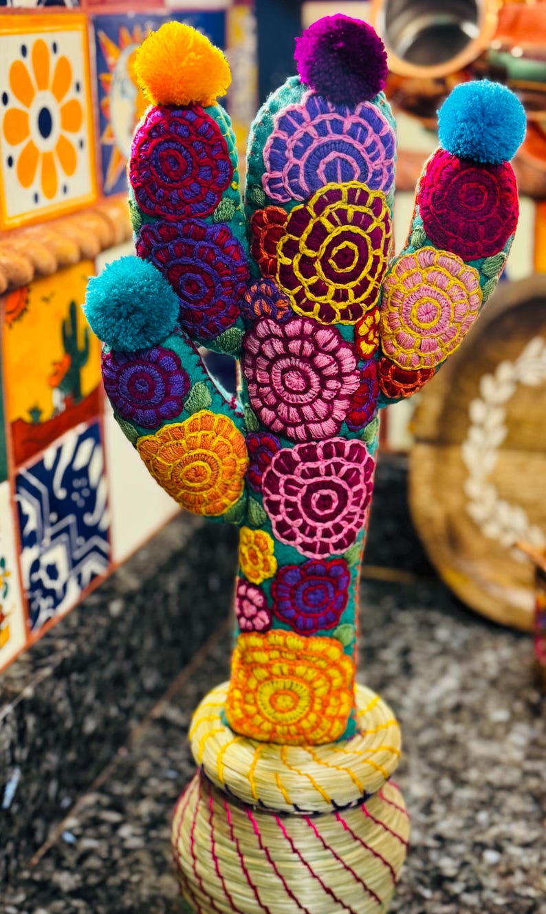 Op de afbeelding: Een kleurrijke geborduurde cactus met pompoenen erop, staand in een geweven pot. De cactus is gemaakt van stof en heeft een verscheidenheid aan felle kleuren, waaronder rood, roze, geel, blauw en groen.