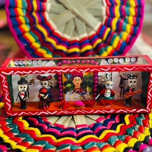 May include: A vibrant, handcrafted Day of the Dead diorama featuring miniature figures and a portrait of Frida Kahlo. The red-framed box sits atop a colorful woven mat, showcasing a festive and cultural theme.