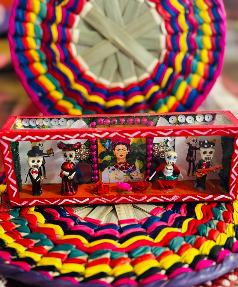 May include: A vibrant, handcrafted Day of the Dead diorama featuring miniature figures and a portrait of Frida Kahlo. The red-framed box sits atop a colorful woven mat, showcasing a festive and cultural theme.