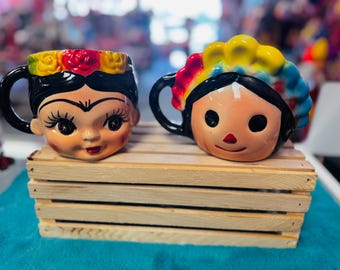 May include: Two colorful ceramic mugs with painted faces. The mugs are decorated with flowers and a colorful design. The mugs are sitting on a wooden crate.