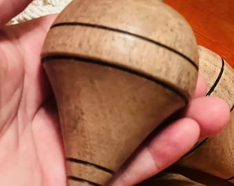 May include: A light brown wooden spinning top with a smooth, rounded body and a pointed base. The toy features two thin, dark horizontal lines etched around the upper section and two near the bottom, with a small metal tip at the very end.