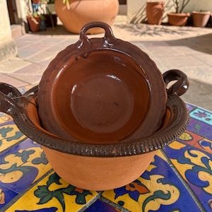 May include: Two terracotta clay bowls, one nested inside the other, with a dark brown rim and handles. The bowls are sitting on a colorful, patterned tile surface. The bowls are likely for cooking or serving.