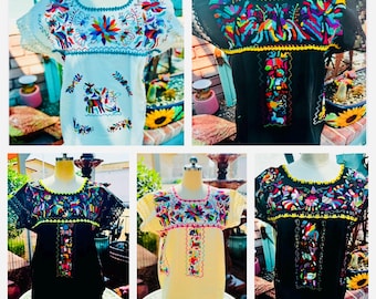 May include: Collection of embroidered blouses in white, black, and yellow. Each blouse features colorful floral and animal designs around the neckline and sleeves. The blouses are displayed on mannequins, showcasing the intricate embroidery.