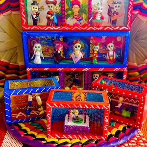 May include: A colorful Day of the Dead folk art display featuring multiple shadow boxes. The boxes are decorated with vibrant red, blue, and gold paint, and contain miniature skeleton figures in various outfits. The display sits on a woven, multi-colored base.
