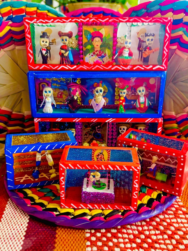 May include: A colorful Day of the Dead folk art display featuring multiple shadow boxes. The boxes are decorated with vibrant red, blue, and gold paint, and contain miniature skeleton figures in various outfits. The display sits on a woven, multi-colored base.