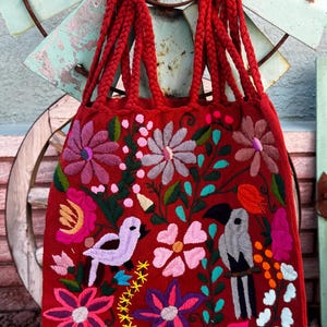 May include: A red tote bag with braided handles and intricate floral and bird embroidery. The embroidered design features pink, purple, and white flowers, along with a light purple bird and a gray and black bird. The bag is a vibrant accessory.