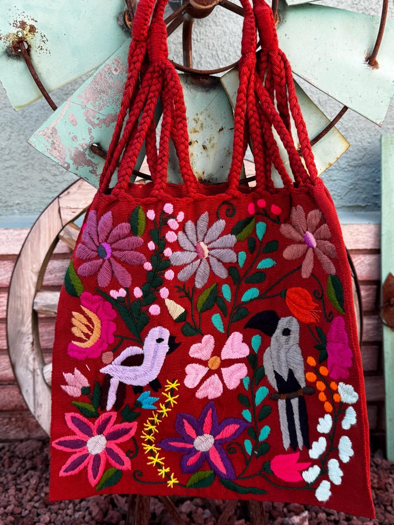 May include: A red tote bag with braided handles and intricate floral and bird embroidery. The embroidered design features pink, purple, and white flowers, along with a light purple bird and a gray and black bird. The bag is a vibrant accessory.