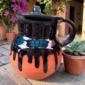 May include: A handmade, terracotta pitcher with a black glazed top and handle. The black glaze drips down the sides, contrasting with the orange-brown clay. The pitcher has a lid and a curved spout.