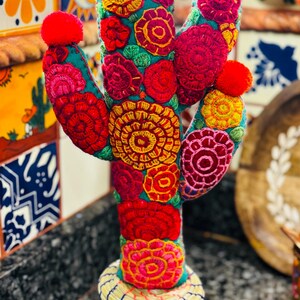 Op de afbeelding: Een kleurrijke geborduurde cactus met roze pompoenen bovenop, staand in een geweven mand. De cactus is versierd met rode, gele, groene en roze bloemen.