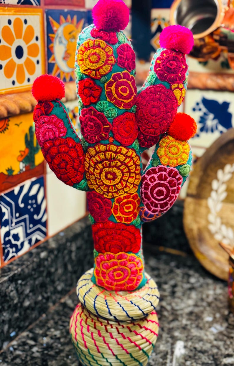 Op de afbeelding: Een kleurrijke geborduurde cactus met roze pompoenen bovenop, staand in een geweven mand. De cactus is versierd met rode, gele, groene en roze bloemen.