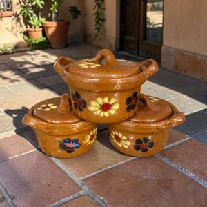 May include: Three small, brown ceramic pots with lids, stacked on a brick surface. Each pot is decorated with a floral design in yellow, red, and black. The pots have handles on the sides and the lids.