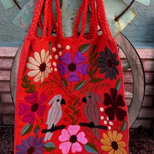 May include: A vibrant red tote bag with braided handles, adorned with colorful embroidered flowers and two birds perched on a branch. The bag's design features a variety of floral patterns in shades of purple, pink, yellow, and white.
