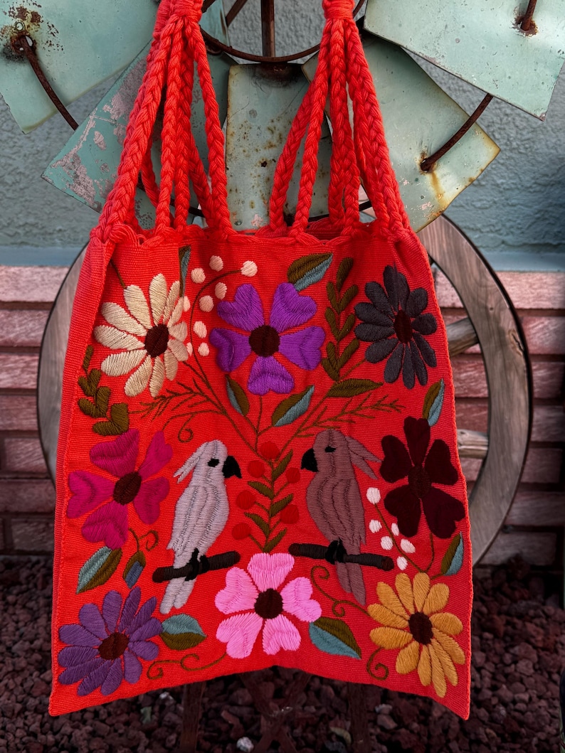 May include: A vibrant red tote bag with braided handles, adorned with colorful embroidered flowers and two birds perched on a branch. The bag's design features a variety of floral patterns in shades of purple, pink, yellow, and white.