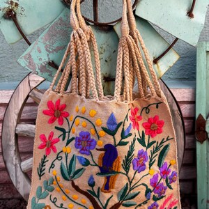 May include: Beige tote bag with embroidered floral and animal designs. The bag features a bird perched on a tree branch, surrounded by pink, purple, and blue flowers. The handles are made of braided beige rope.