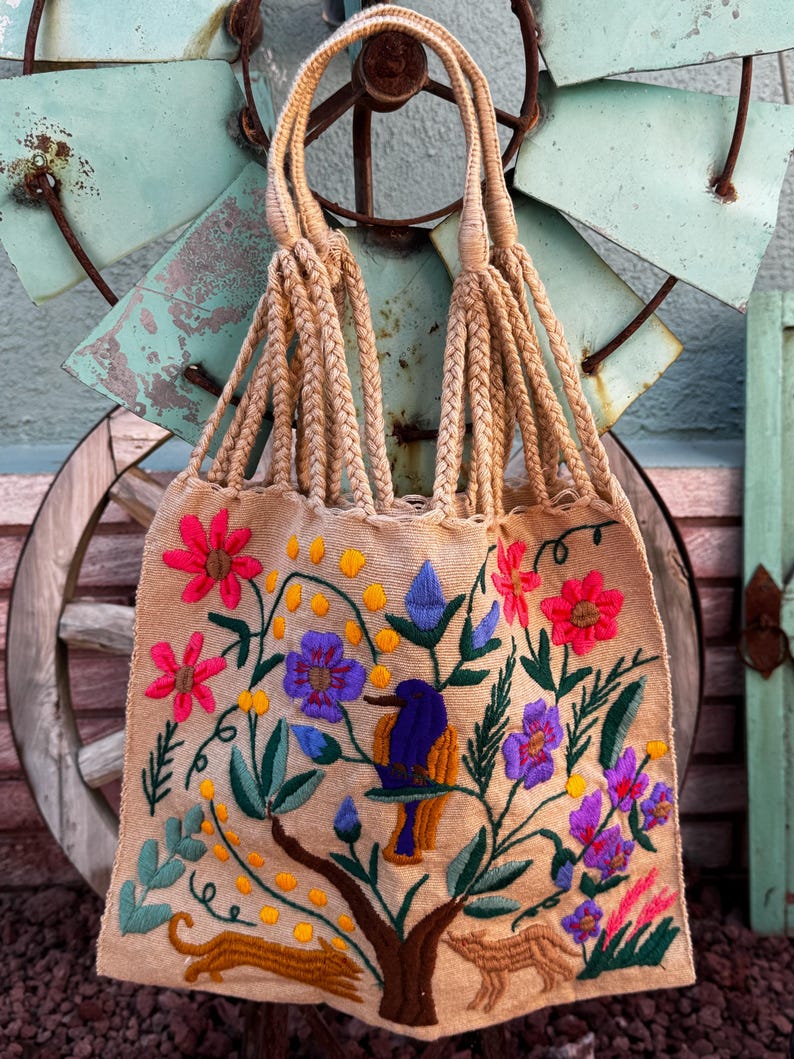 May include: Beige tote bag with embroidered floral and animal designs. The bag features a bird perched on a tree branch, surrounded by pink, purple, and blue flowers. The handles are made of braided beige rope.