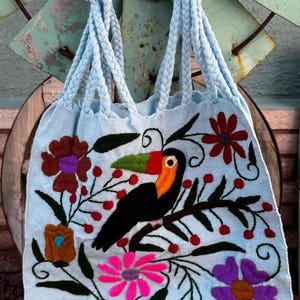 May include: Light blue tote bag with braided handles, featuring an embroidered toucan perched on a branch with flowers and berries. The bag's design includes pink, purple, brown, and red floral accents, creating a vibrant, handcrafted look.