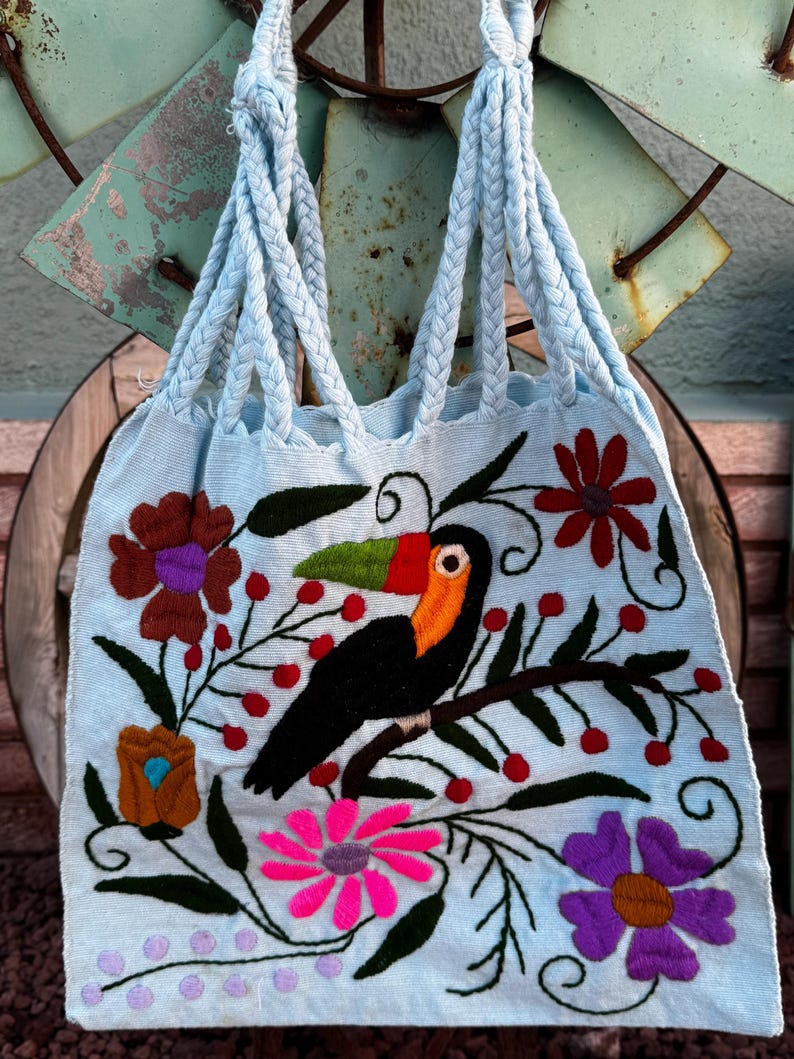 May include: Light blue tote bag with braided handles, featuring an embroidered toucan perched on a branch with flowers and berries. The bag's design includes pink, purple, brown, and red floral accents, creating a vibrant, handcrafted look.