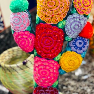 Op de afbeelding: Een kleurrijke geborduurde cactus met een groene basis en meerdere bloemen in tinten roze, rood, geel, paars en blauw. De cactus heeft een pompon bovenop.
