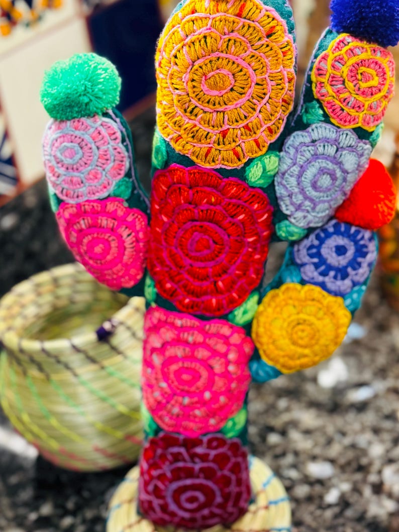 Op de afbeelding: Een kleurrijke geborduurde cactus met een groene basis en meerdere bloemen in tinten roze, rood, geel, paars en blauw. De cactus heeft een pompon bovenop.