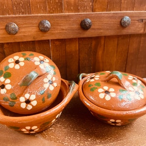 May include: Two terracotta clay bowls with lids. The bowls and lids are decorated with white daisy-like flowers and green leaves. The lids have handles. The bowls are on a wooden surface.