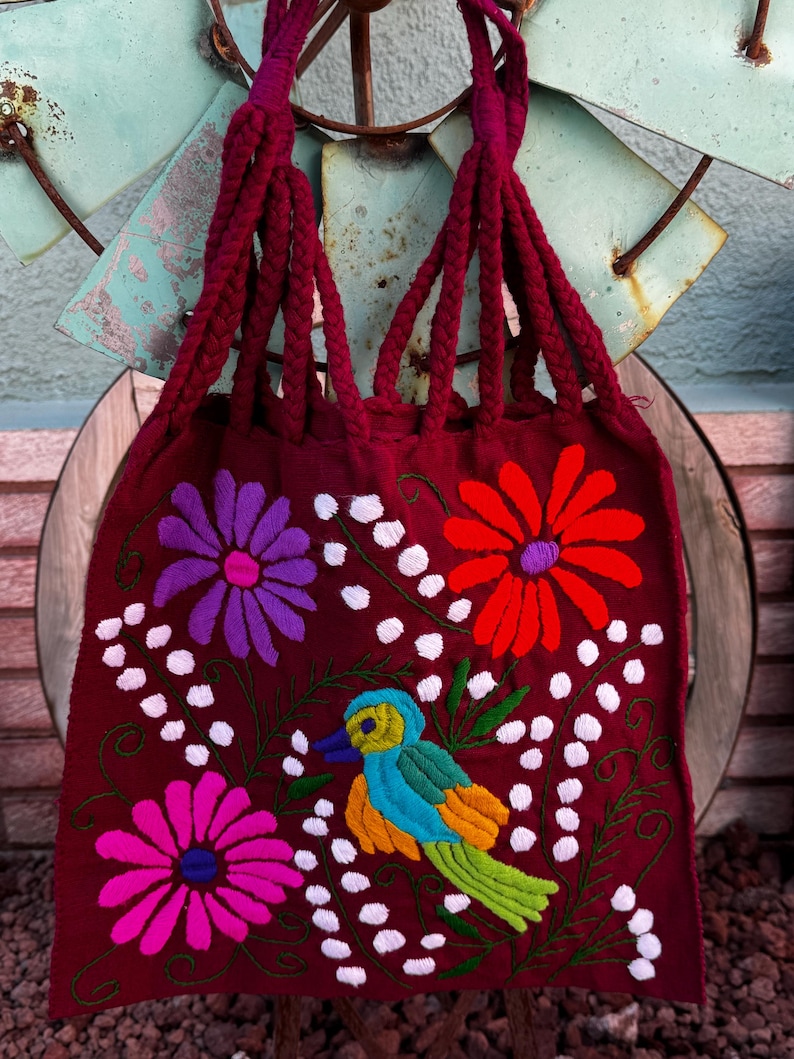 May include: A burgundy tote bag with braided handles and colorful floral and bird embroidery. The embroidered flowers are purple, pink, and red, with a blue, yellow, and green bird. White dots and green vines accent the design.
