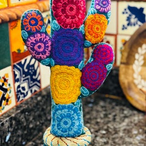 Op de afbeelding: Een kleurrijke geborduurde cactus met een groene basis en een gele pompon bovenop. De cactus is versierd met meerkleurige bloemen in tinten roze, rood, oranje, geel, blauw en paars. De cactus staat in een geweven mand.