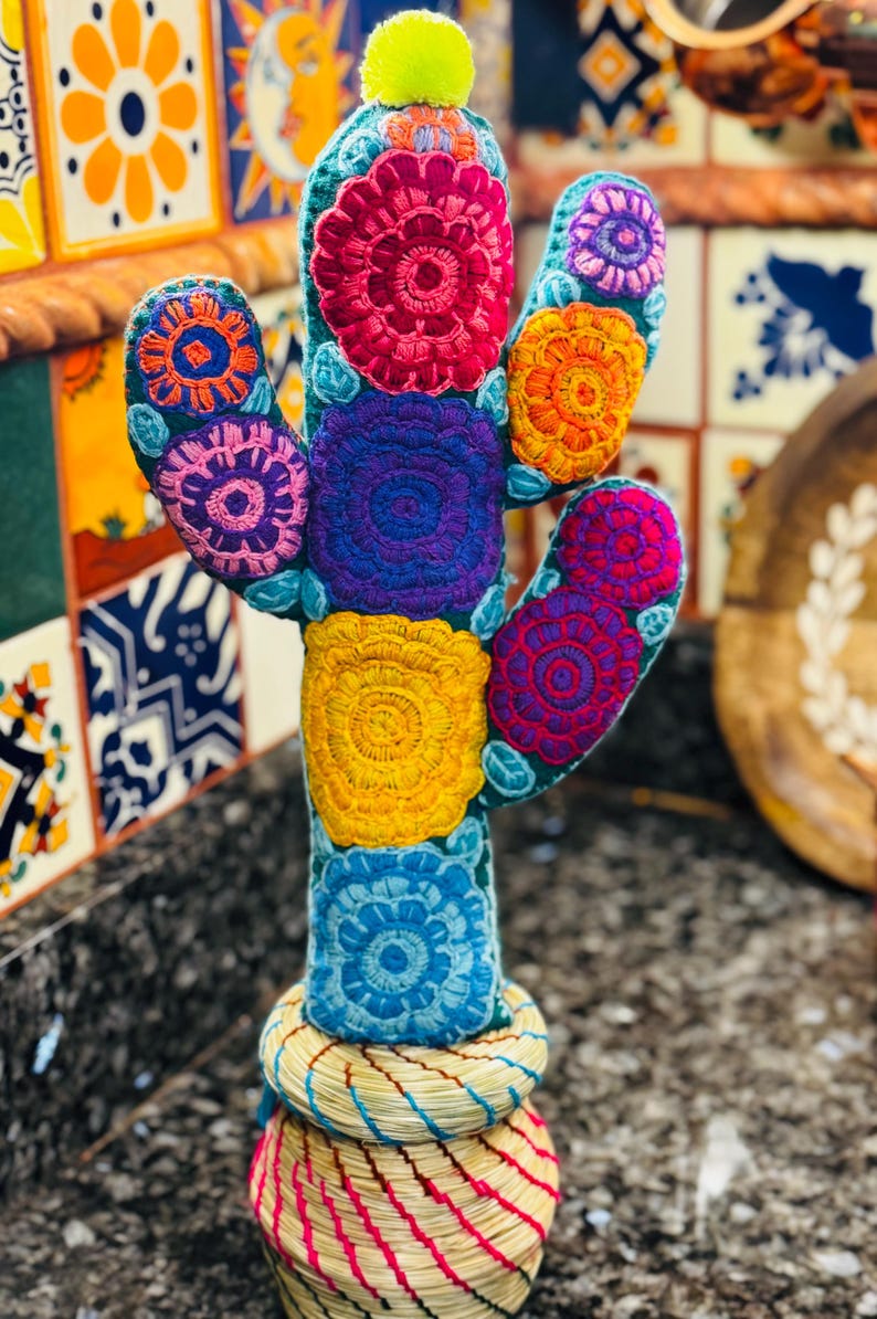Op de afbeelding: Een kleurrijke geborduurde cactus met een groene basis en een gele pompon bovenop. De cactus is versierd met meerkleurige bloemen in tinten roze, rood, oranje, geel, blauw en paars. De cactus staat in een geweven mand.