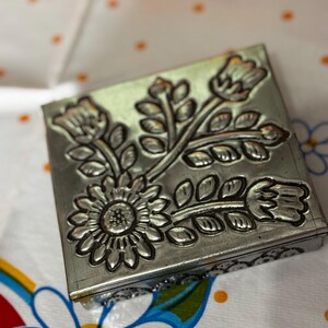 May include: A square, silver-colored metal box with a floral design. The embossed flowers include a sunflower and other stylized blooms and leaves. The box is resting on a white tablecloth with a floral pattern.