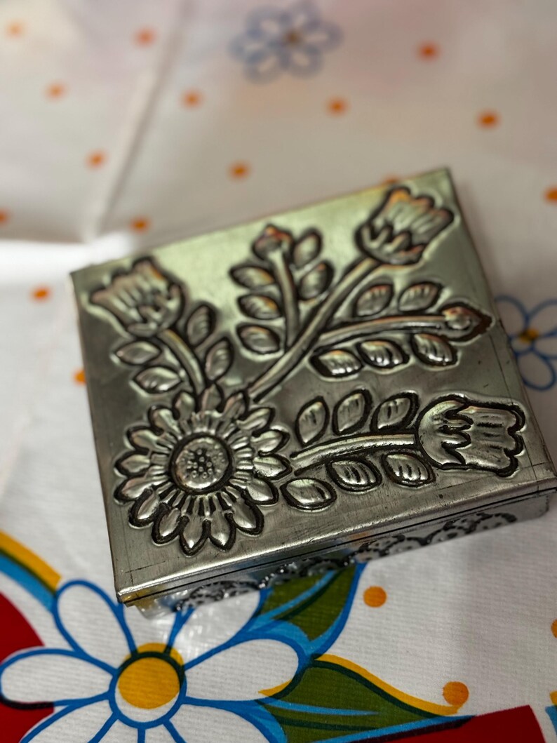 May include: A square, silver-colored metal box with a floral design. The embossed flowers include a sunflower and other stylized blooms and leaves. The box is resting on a white tablecloth with a floral pattern.