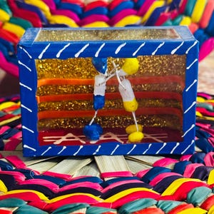 May include: A blue box with a clear top and white diagonal lines on the edges. Inside, there are gold glitter and colorful horizontal stripes. Two small figures hang inside the box. The box sits on a colorful woven mat.