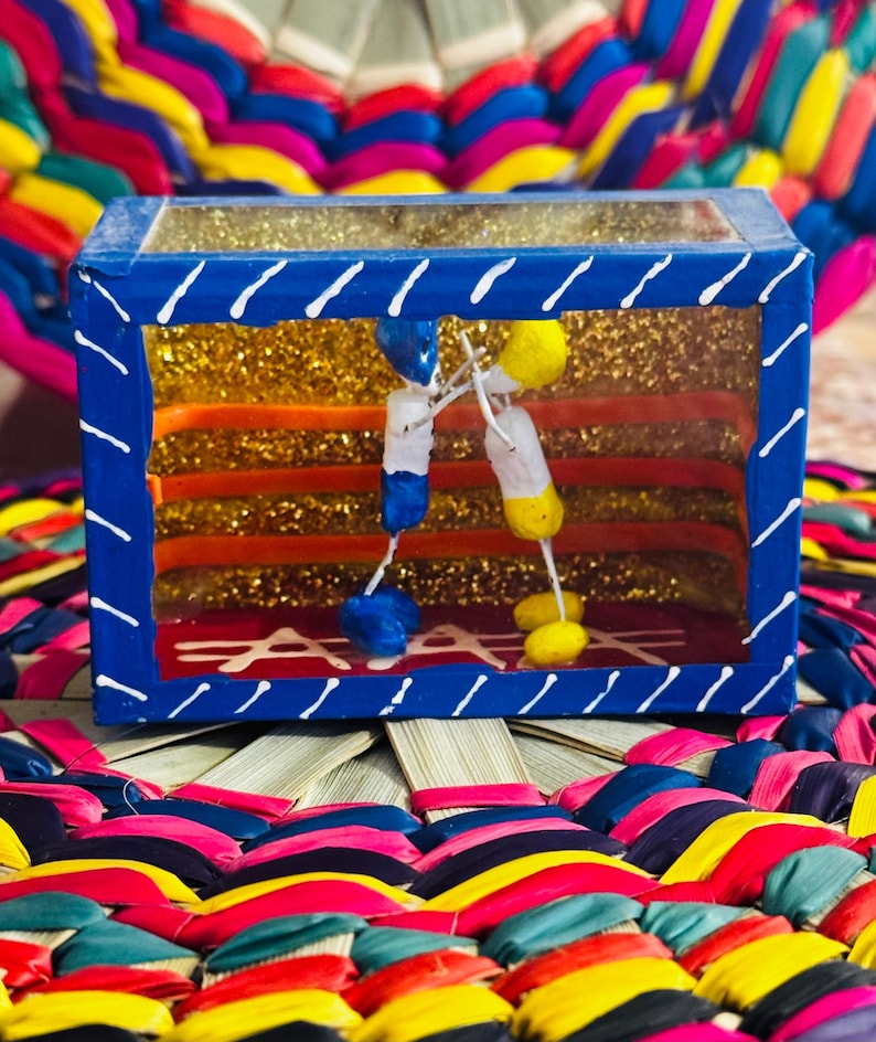 May include: A blue box with a clear top and white diagonal lines on the edges. Inside, there are gold glitter and colorful horizontal stripes. Two small figures hang inside the box. The box sits on a colorful woven mat.