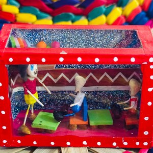 May include: A handcrafted, red wooden diorama with a clear top, featuring miniature figures and furniture inside. The diorama has a red frame with white polka dots. The background is a colorful woven pattern.