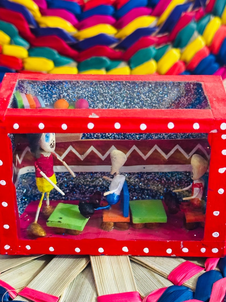 May include: A handcrafted, red wooden diorama with a clear top, featuring miniature figures and furniture inside. The diorama has a red frame with white polka dots. The background is a colorful woven pattern.