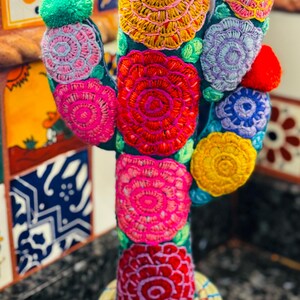 Op de afbeelding: Een kleurrijke geborduurde cactus met een geweven pot. De cactus is gemaakt van stof en heeft een verscheidenheid aan bloemen in tinten roze, rood, geel, blauw en groen. De pot is gemaakt van natuurlijke materialen en heeft een lichtbruine kleur.