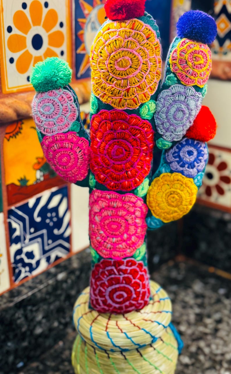 Op de afbeelding: Een kleurrijke geborduurde cactus met een geweven pot. De cactus is gemaakt van stof en heeft een verscheidenheid aan bloemen in tinten roze, rood, geel, blauw en groen. De pot is gemaakt van natuurlijke materialen en heeft een lichtbruine kleur.