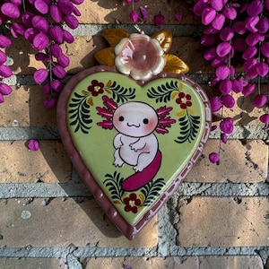 May include: A heart-shaped ceramic wall plaque featuring a light green center with a hand-painted illustration of a white axolotl with pink gills and a tail. The heart is framed by a dark pink border and topped with a sculpted ceramic flower.