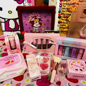 May include: A collection of pink and white beauty products, including lip gloss sets, facial wipes, and a makeup brush. The products are arranged on a pink strawberry-patterned surface, with a framed Strawberry Shortcake image in the background.