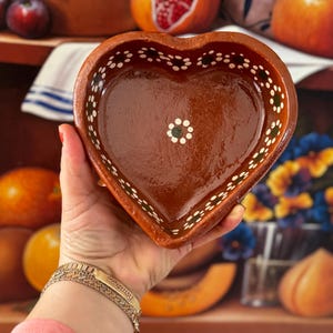 May include: A heart-shaped, brown ceramic bowl with white and green dot detailing around the rim. The bowl is held in a hand, with a gold bracelet visible. The background includes fruit and other decorative items.