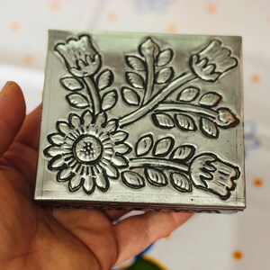 May include: A square, silver-colored metal box with a floral design. The box features an embossed pattern of a large sunflower and several stylized flowers and leaves. The box is held in a hand, and the background is a patterned fabric.