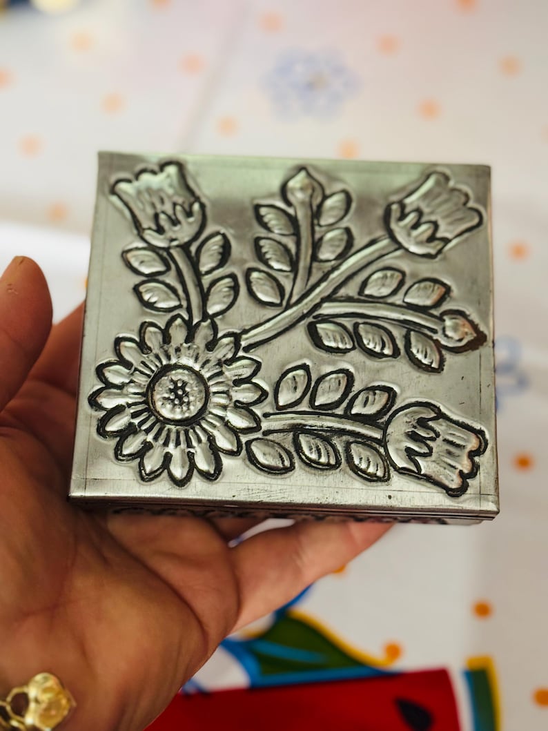 May include: A square, silver-colored metal box with a floral design. The box features an embossed pattern of a large sunflower and several stylized flowers and leaves. The box is held in a hand, and the background is a patterned fabric.