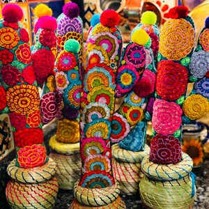 Op de afbeelding: Een groep kleurrijke, geborduurde cactusfiguren met pompoenen erop. De cactussen zijn gemaakt van stof en hebben verschillende bloemenpatronen in tinten rood, roze, geel, blauw en groen. De cactussen staan op geweven manden.