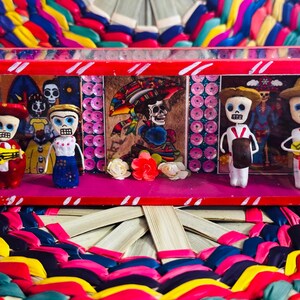 May include: A Day of the Dead themed display featuring miniature skeleton figures in a red and pink box. The figures are dressed in colorful outfits and play musical instruments. The background includes painted images and decorative elements.