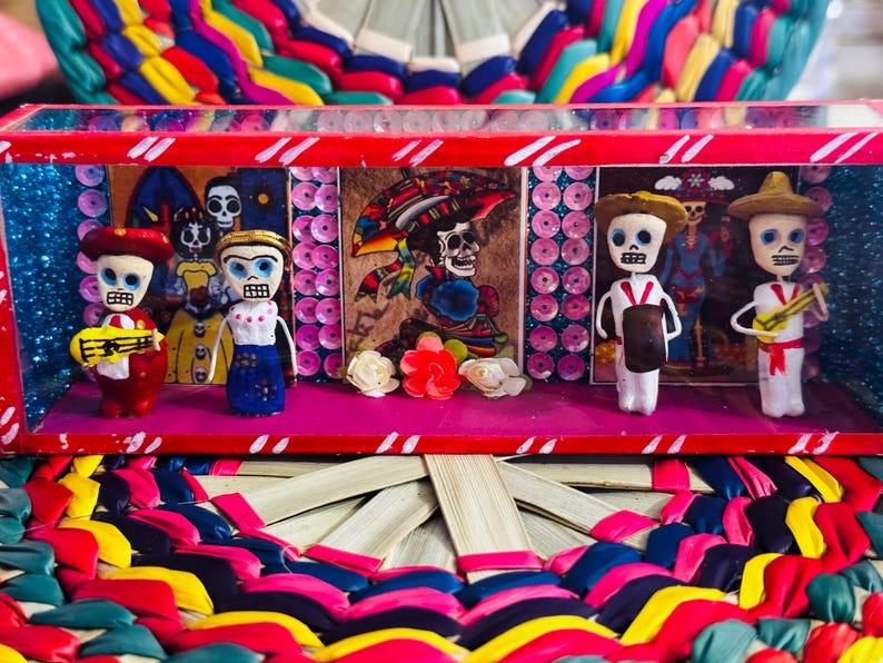 May include: A Day of the Dead themed display featuring miniature skeleton figures in a red and pink box. The figures are dressed in colorful outfits and play musical instruments. The background includes painted images and decorative elements.