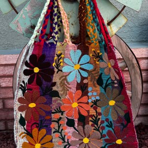 May include: A colorful, embroidered tote bag with a floral design. The bag features large, stylized flowers in shades of brown, orange, blue, and yellow, set against vertical stripes of various colors. The handles are braided in multiple colors.