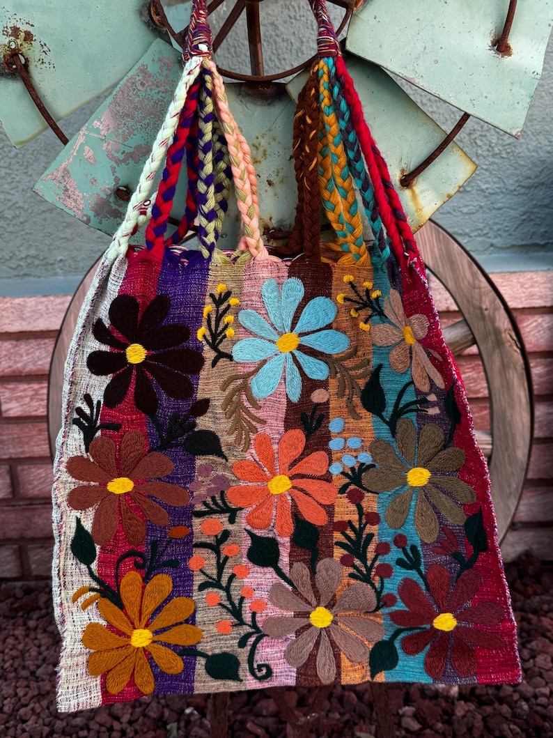 May include: A colorful, embroidered tote bag with a floral design. The bag features large, stylized flowers in shades of brown, orange, blue, and yellow, set against vertical stripes of various colors. The handles are braided in multiple colors.